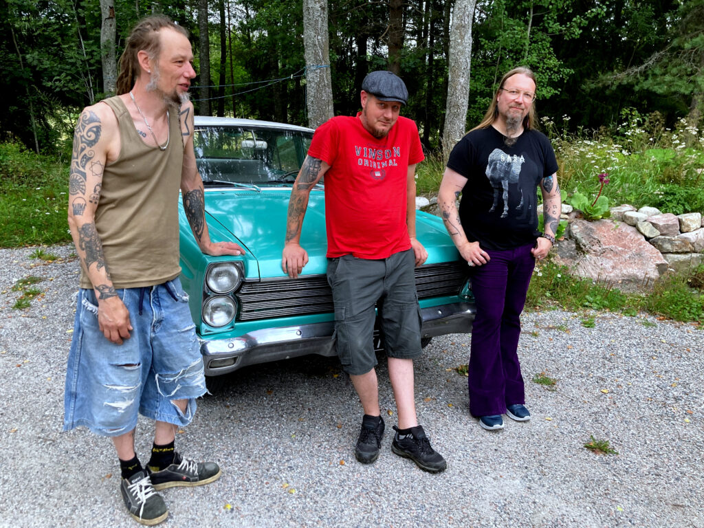 Photo of the band in front of a turquoise car. From left to right, Jonesi, Osku, and Tuomo